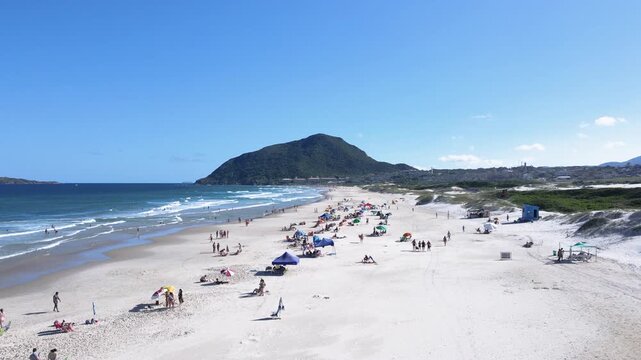 Wide aerial view of Santinho Beach in Florianopolis, Brazil, showing people relaxing on white sand, ocean waves and the Morro das Aranhas mountain under a clear blue sky.
