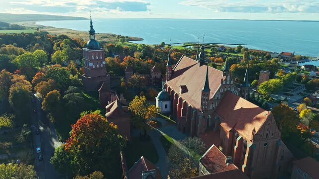 Aerial drone view of the castle and cathedral complex in Frombork, Poland surrounded by vibrant autumn forest. Historic brick architecture and towers dominate the skyline in golden seasonal light.