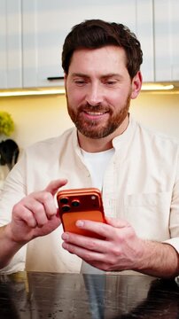 Middle-aged man smiling while typing on smartphone in home kitchen beside fresh fruit juice and blender. Young boy checks quick recipe tips for diet goals often on screen enjoying calm mood indoors