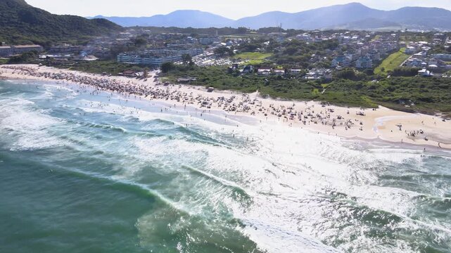 Cinematic orbital pull-back aerial shot of Santinho Beach in Florianopolis, Brazil, revealing a crowded shoreline, turquoise ocean waves and the Morro das Aranhas mountain.
