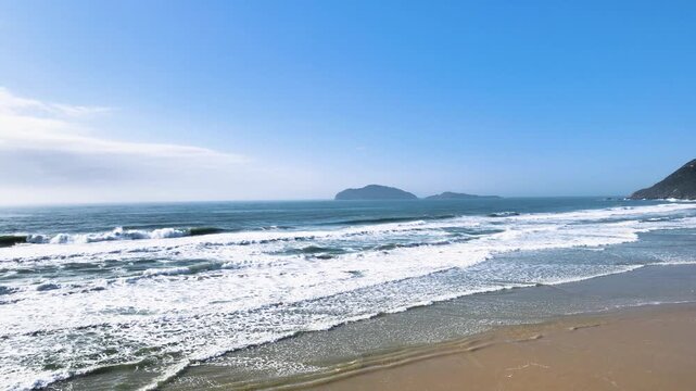 Low-altitude aerial view of turquoise ocean waves crashing on the shore of Santinho Beach in Florianopolis, Brazil, with islands on the horizon under a clear blue sky.
