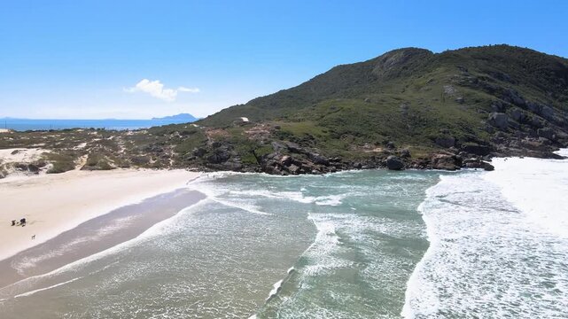 Low-altitude aerial wide shot of Santinho Beach in Florianopolis, Brazil, showing turquoise waves crashing on the shore with the Morro dos Ingleses mountain under a clear blue sky.
