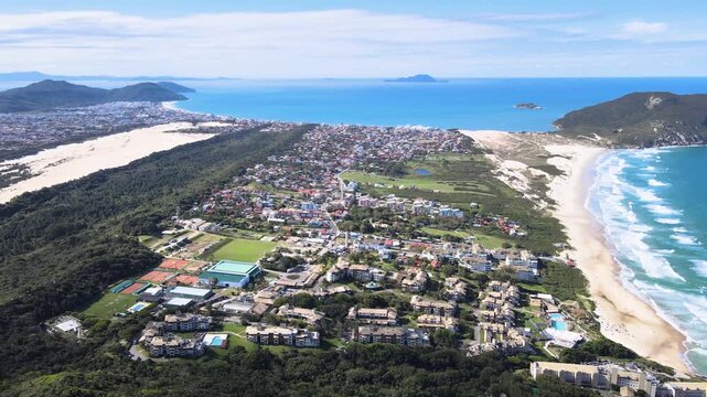 High-altitude aerial panorama of Santinho and Ingleses beaches in Florianopolis, Brazil, showcasing white sand dunes, coastal neighborhoods and the Atlantic Ocean under a bright sky.
