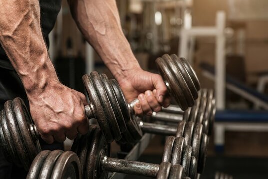 A close-up shot of a muscular man's strong, veiny hands grabbing heavy dumbbells from a rack in a gym.