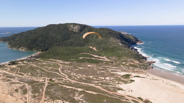 Aerial wide shot of a paramotor flying over the Santinho Beach sand dunes in Florianopolis, Brazil, with Morro dos Ingleses mountain and turquoise ocean in the background.
