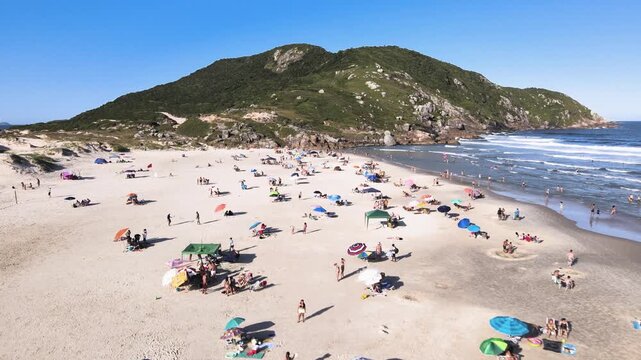 Low-altitude aerial wide shot of Santinho Beach in Florianopolis, Brazil, featuring crowds of tourists on the sand and the Morro dos Ingleses mountain under a bright sunny sky.
