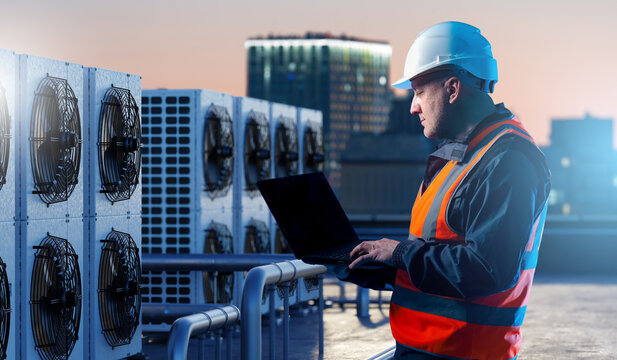 Building maintenance, HVAC monitoring. Technician in safety helmet uses laptop on rooftop to check air conditioning systems, analyzing performance and ensuring reliable cooling operation.
