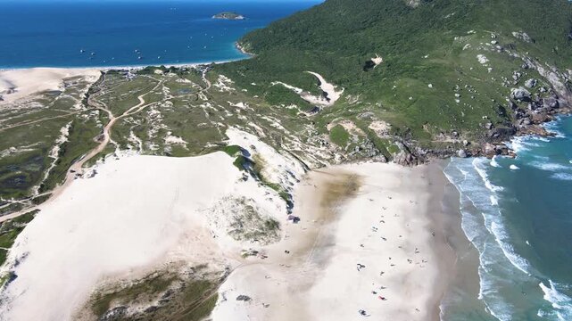 Cinematic aerial wide panorama of Santinho Beach dunes and Morro dos Ingleses in Florianopolis, Brazil, featuring turquoise ocean waves, white sand and coastal vegetation on a sunny day.
