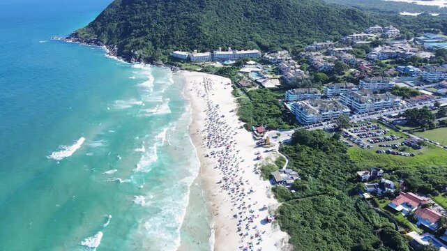 High angle aerial view of Santinho Beach in Florianopolis, Brazil, showing crowded shore, seaside apartments, parking lots and the green Morro das Aranhas mountain on a sunny day
