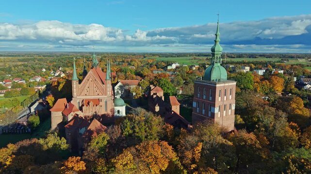 Aerial drone view of the castle and cathedral complex in Frombork, Poland surrounded by vibrant autumn forest. Historic brick architecture and towers dominate the skyline in golden seasonal light.