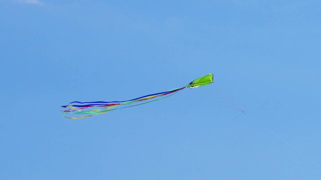 Green kite flying and drifting in clear blue sky with colorful ribbons fluttering in wind on sunny spring day, Family activity, Outdoor leisure
