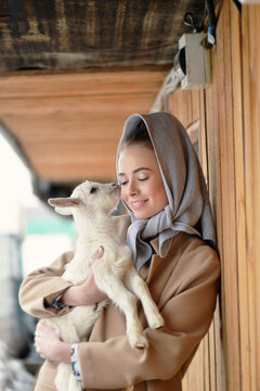 A young woman in a basic coat holds a small goat in her arms. 