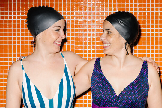 Two joyful women in swim caps against a tiled wall