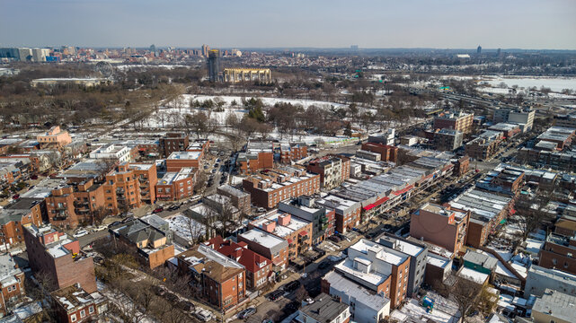 Aerial view showing residential and commercial buildings with snow covered rooftops lining streets next to a large urban park under a clear sky in corona, queens, new york