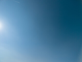 Obraz premium Expansive view of a bright clear blue sky, featuring a glowing sun flare in the corner and a faint airplane contrail tracing across the upper backdrop, offering copy space for various designs
