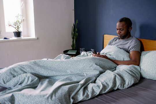 Adult black man using smartphone lying on comfortable bed in bedroom