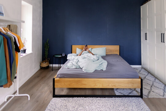 Distance view of man using smartphone lying on bed in bedroom at home