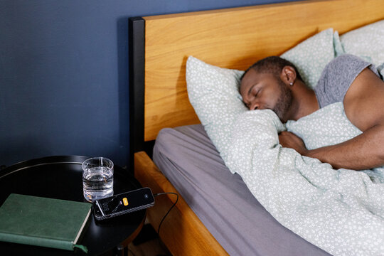 Adult black man sleeping in bed near table in bedroom at home