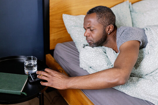 Man turning off alarm on phone on table while lying on bed in bedroom