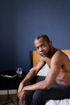 Serious adult black man with naked torso on bed and looking at camera