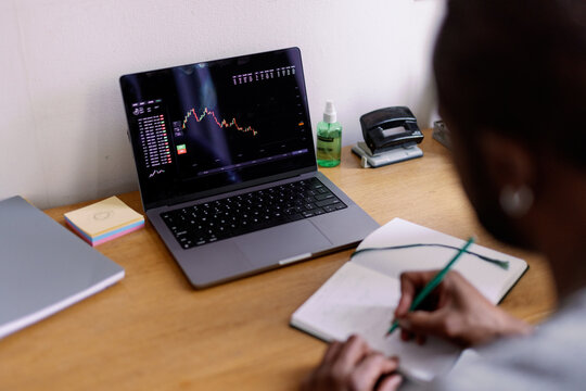 Man watching stock market graph on laptop and writing in notebook