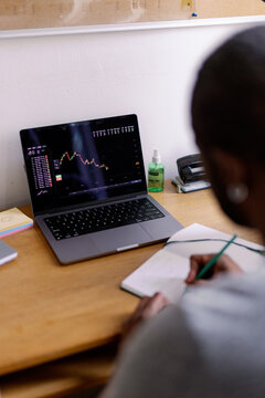 Partial man watching market graph on laptop and writing in notebook