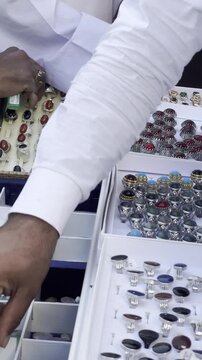 Explore a vibrant display of colorful loose gemstones and traditional men's rings at Bilal Market. Watch shoppers browse through trays of beautiful polished stones in bustling local jewelry bazaar.