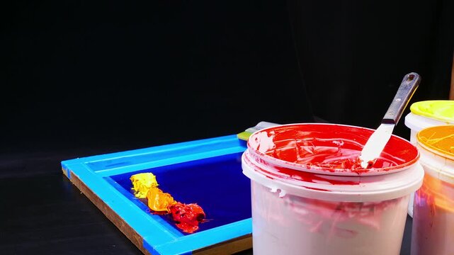 A bright studio setup featuring open buckets of vibrant inks beside a blue screen printing frame. The mix of tools and hues reflects the artistry and preparation behind professional printmaking.