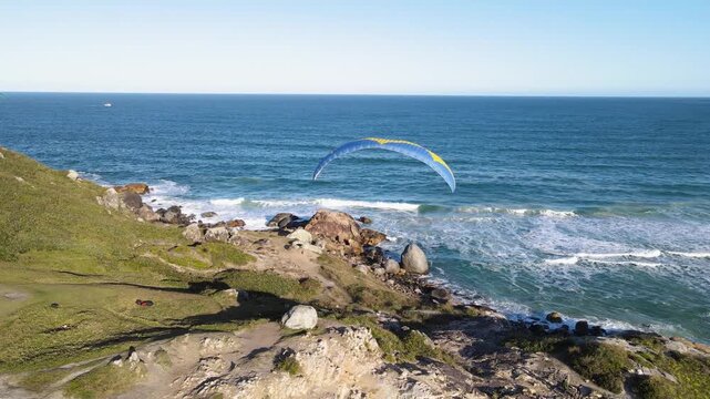 Cinematic aerial orbital shot of paragliders flying over the rocky cliffs of Morro dos Ingleses in Florianopolis, Brazil, with Santinho Beach and the Atlantic Ocean in the background.
