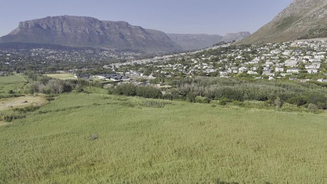 Drone flies over grassy plains near river toward the Imizamo Yethu informal settlement on a sunny morning in Hout Bay, Cape Town, South Africa