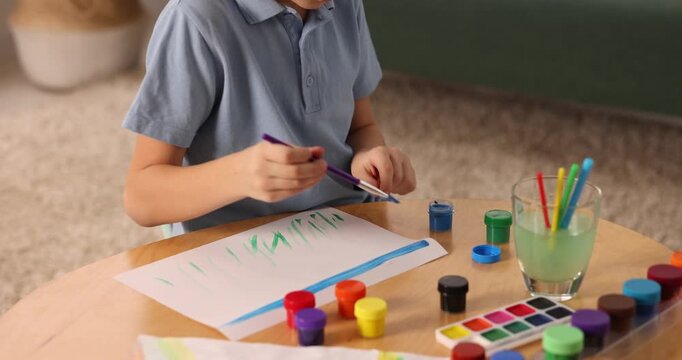 Little boy drawing sky with brush and paints at wooden table indoors. Camera moving forward