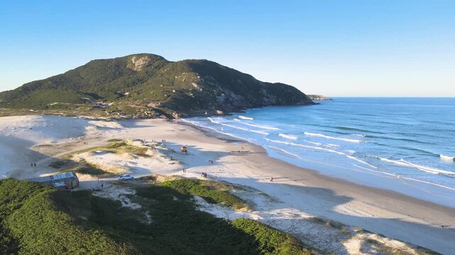 Cinematic aerial orbital shot of Santinho Beach in Florianopolis, Brazil, revealing white sand dunes, turquoise ocean waves and the Morro dos Ingleses mountain on a sunny day.
