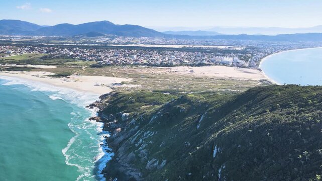 Cinematic aerial panorama of Santinho Beach in Florianopolis, Brazil, featuring turquoise ocean waves crashing on rocky cliffs, white sand dunes and Morro dos Ingleses mountain on a sunny day.
