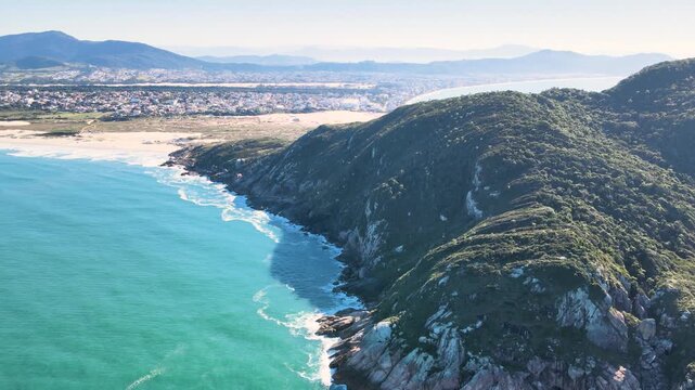 Cinematic aerial panorama of Santinho Beach in Florianopolis, Brazil, featuring turquoise ocean waves crashing on rocky cliffs, white sand dunes and Morro dos Ingleses mountain on a sunny day.
