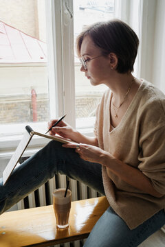 A woman with coffee sitting next to window and writing notes