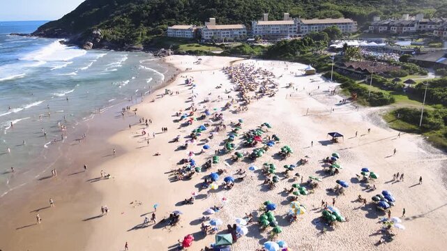 Low-to-high aerial tilt-up shot of Santinho Beach in Florianopolis, Brazil, revealing a crowded summer shore and the luxury Costao do Santinho Resort at the foot of Morro das Aranhas
