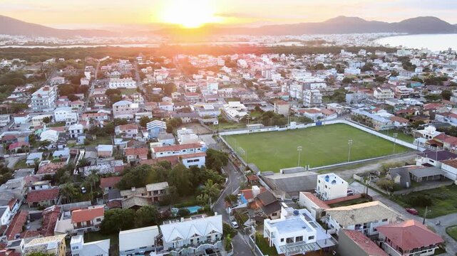 Cinematic aerial orbital hyperlapse of Santinho neighborhood in Florianopolis, Brazil, at sunset, showing the Nautico soccer field and coastal houses under a fast-moving golden sky.
