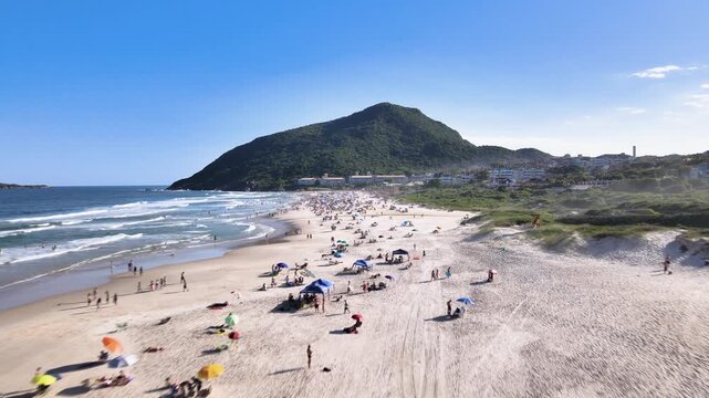 Fast low-altitude aerial drone shot flying over Santinho Beach in Florianopolis, Brazil, towards Morro das Aranhas mountain with crowds of people and ocean waves.
