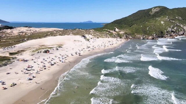 Cinematic aerial orbital shot of Santinho Beach in Florianopolis, Brazil, revealing white sand dunes, turquoise ocean waves and the Morro dos Ingleses mountain on a sunny day.
