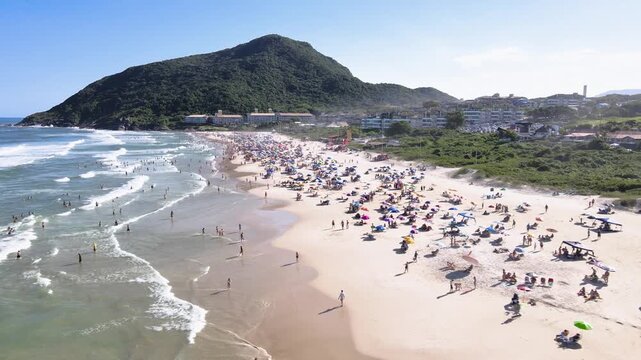 Low-altitude aerial wide shot of a crowded Santinho Beach in Florianopolis, Brazil, featuring tourists, colorful umbrellas and the green Morro das Aranhas mountain on a sunny day.

