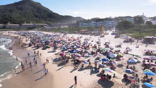 Low-altitude aerial view of a crowded Santinho Beach in Florianopolis, Brazil, with colorful umbrellas, people enjoying summer and Morro das Aranhas mountain on a sunny day.
