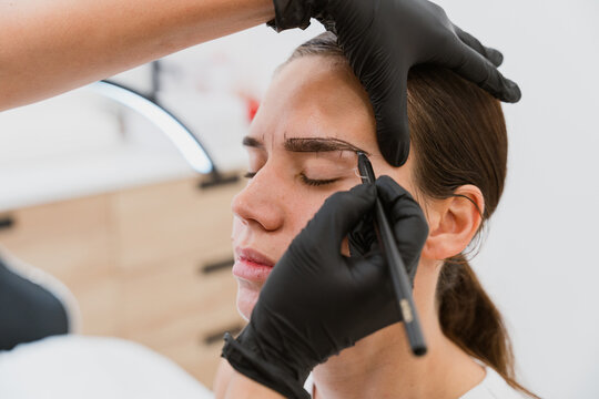 Beautician drawing eyebrow shape on young woman face with pencil