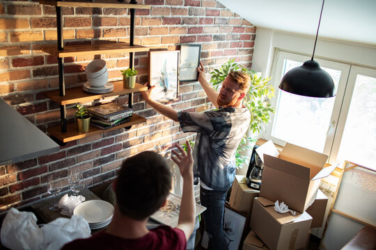 Two men hang framed art in brick-walled apartment