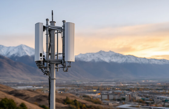 Modern telecommunications cell tower mast with antennas standing against beautiful snowy mountains and city valley during sunset