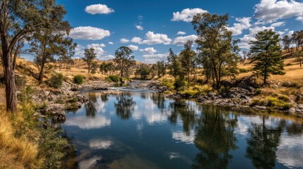 Obraz premium Serene River Scene with Reflective Water Surrounded by Lush Trees and Golden Hills Under a Bright Blue Sky with Fluffy White Clouds