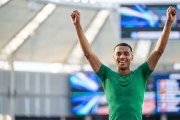 A young male athlete in a green shirt raises his arms in victory inside a stadium