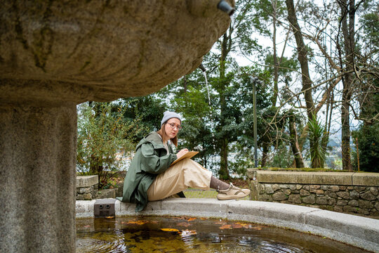 Woman sketching on a notepad while sitting by a stone fountain.