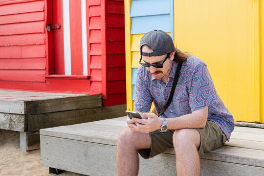 Man sits relaxed near red and yellow beach huts in Middle Brighton