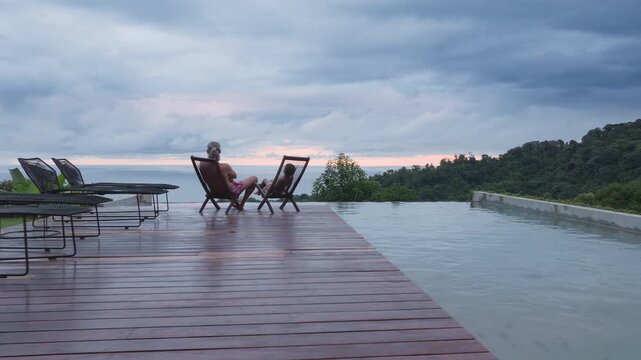 In a tranquil scene, a father and his son spend quality time unwinding at their beautiful home, with a stunning view of a scenic swimming pool and lush landscapes in picturesque Costa Rica
