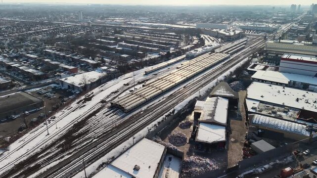 High angle aerial perspective of a commuter train yard and railway depot surrounded by an industrial and residential urban area during a sunny winter day, with snow covering the landscape